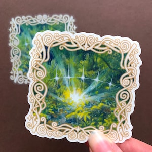 May include: Two stickers featuring a forest scene with a bright, sunlit opening. The artwork is framed with a gold, ornate border. The stickers have a white background and are held by a hand.