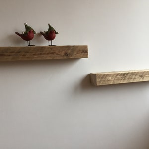 May include: Two rustic wooden shelves mounted on a white wall. The top shelf has two small red bird figurines with green hats.