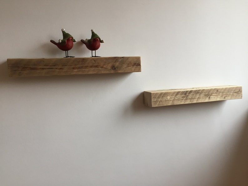 May include: Two rustic wooden shelves mounted on a white wall. The top shelf has two small red bird figurines with green hats.