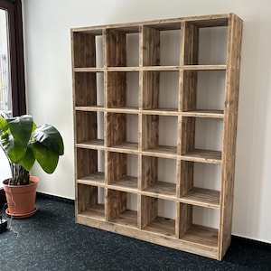 Könnte beinhalten: Ein großes, rustikales Bücherregal aus Holz mit mehreren quadratischen Fächern. Das Bücherregal ist aus hellbraunem Holz gefertigt und steht vor einer weißen Wand. Links vom Bücherregal ist eine Topfpflanze zu sehen.