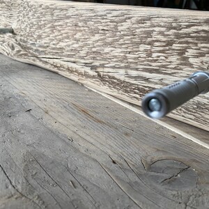 May include: A close-up of a wooden plank with two silver screws being inserted into the wood. The screws are being driven in by a power screwdriver.