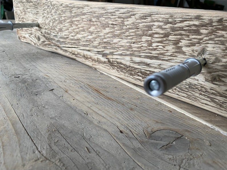 May include: A close-up of a wooden plank with two silver screws being inserted into the wood. The screws are being driven in by a power screwdriver.