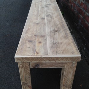 Puede incluir: Un banco de madera r&uacute;stico con un acabado natural. El banco tiene un dise&ntilde;o sencillo con una parte superior rectangular y dos patas a cada lado. La madera tiene un aspecto desgastado con nudos y patrones de veta.