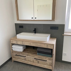 May include: A wooden bathroom vanity with a white porcelain sink and two drawers. The vanity has a shelf above the drawers and a mirror cabinet above the sink. The vanity is in a bathroom with grey tiled walls.