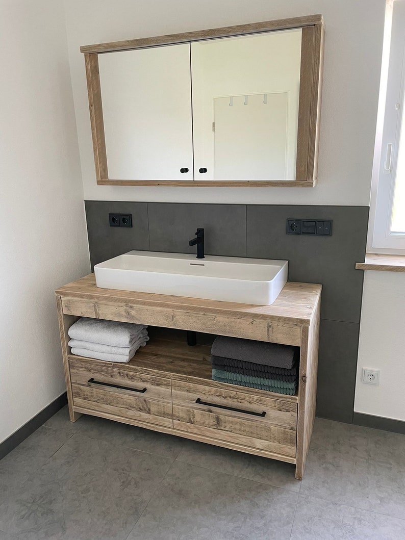 May include: A wooden bathroom vanity with a white porcelain sink and two drawers. The vanity has a shelf above the drawers and a mirror cabinet above the sink. The vanity is in a bathroom with grey tiled walls.