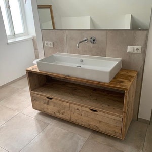 May include: A white porcelain bathroom sink with a wooden vanity cabinet with two drawers. The vanity is made of reclaimed wood with a rustic finish.