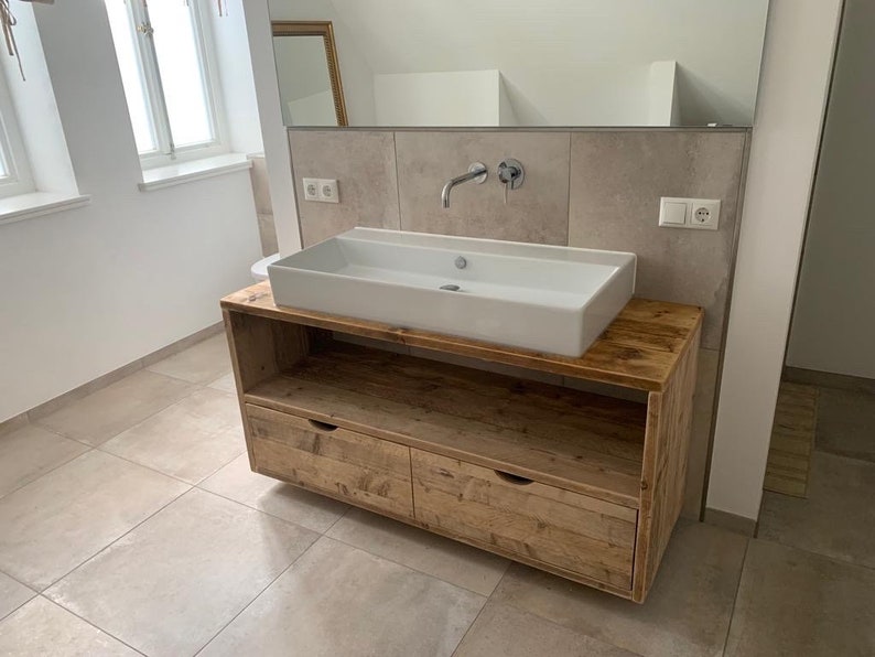 May include: A white porcelain bathroom sink with a wooden vanity cabinet with two drawers. The vanity is made of reclaimed wood with a rustic finish.
