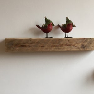 May include: Two red birds wearing green hats with white pom-poms are perched on a rustic wooden shelf.