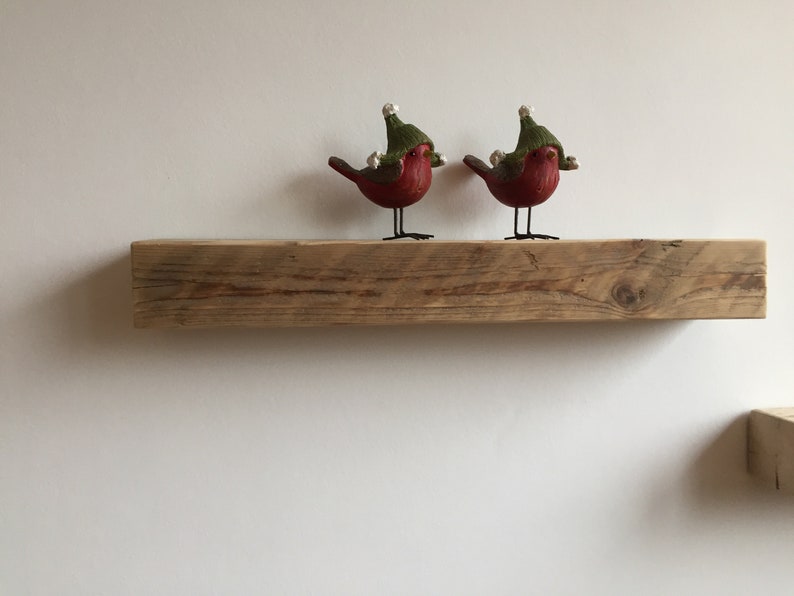 May include: Two red birds wearing green hats with white pom-poms are perched on a rustic wooden shelf.
