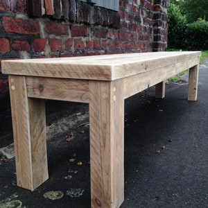 Puede incluir: Un banco de madera r&uacute;stico con un acabado de madera natural. El banco tiene un dise&ntilde;o sencillo con cuatro patas y una parte superior rectangular. El banco est&aacute; hecho de madera recuperada y tiene un aspecto desgastado.