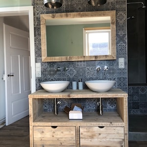 May include: A double vanity bathroom sink with a wooden countertop and two white porcelain sinks. The countertop is made of reclaimed wood and has a rustic look. The vanity is set against a wall with patterned grey tiles.
