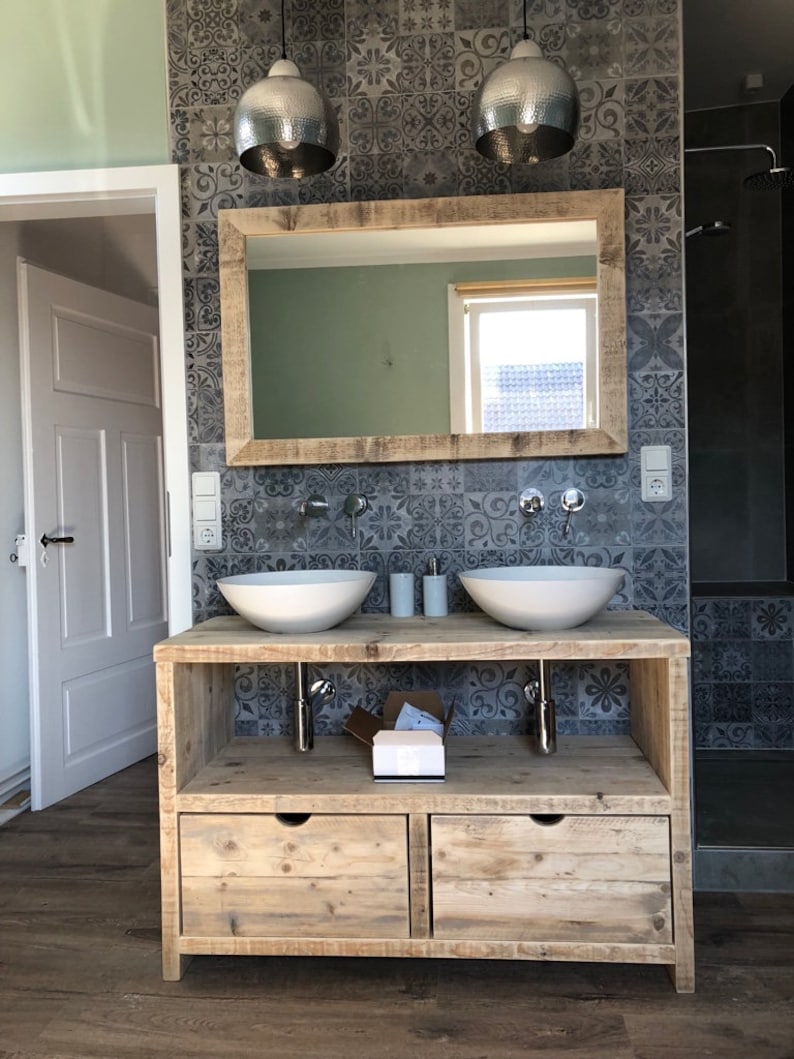 May include: A double vanity bathroom sink with a wooden countertop and two white porcelain sinks. The countertop is made of reclaimed wood and has a rustic look. The vanity is set against a wall with patterned grey tiles.
