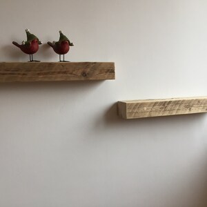 May include: Two rustic wooden shelves mounted on a white wall. The top shelf has two red bird figurines wearing green hats.