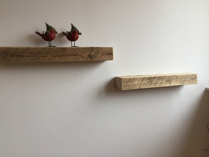 May include: Two rustic wooden shelves mounted on a white wall. The top shelf has two red bird figurines wearing green hats.
