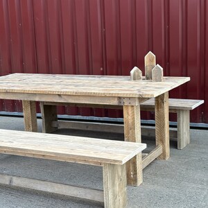 Könnte beinhalten: Ein rustikaler Holz-Picknicktisch mit zwei Bänken. Tisch und Bänke sind aus verwittertem Holz mit natürlicher Oberfläche. Der Tisch ist rechteckig und hat ein schlichtes Design. Die Bänke sind ebenfalls rechteckig und haben ein schlichtes Design. Tisch und Bänke stehen vor einer roten Metallwand.