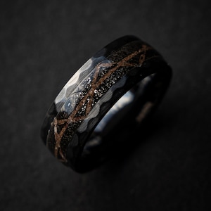 Può includere: Un anello nero e argento con un design testurizzato. L'anello ha una fascia sottile e una parte superiore larga con un motivo di linee e punti.