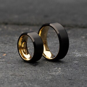 Op de afbeelding: Twee zwarte ringen van wolfraamcarbide met gouden binnenkanten. De ringen zijn op elkaar gestapeld.
