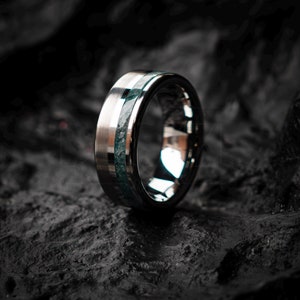 Op de afbeelding: Een zilveren ring met een groene inlay. De ring ligt op een donker, getextureerd oppervlak.