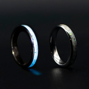 Op de afbeelding: Twee ringen van wolfraamcarbide met een witte inlay. De ring aan de linkerkant gloeit blauw in het donker. De ring aan de rechterkant heeft een witte inlay met een subtiele iriserende glans.