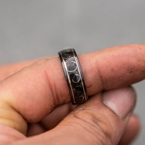 May include: A black and gold ring with a textured design. The ring is worn on a finger.