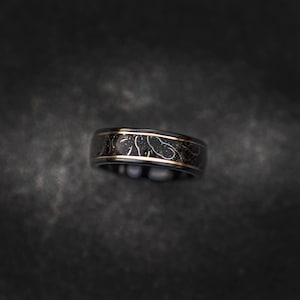 May include: A black ring with a gold band and a white design. The design is a pattern of small circles and lines.