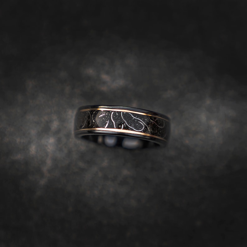 May include: A black ring with a gold band and a white design. The design is a pattern of small circles and lines.