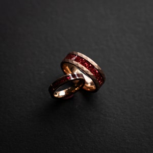 K&ouml;nnte beinhalten: Zwei goldene Eheringe mit einem roten Edelstein-Inlay. Die Ringe sind &uuml;bereinander gestapelt.