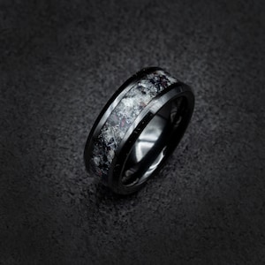 Puede incluir: Un anillo de carburo de tungsteno negro con una incrustación moteada en blanco y negro. El anillo tiene un borde biselado y un acabado pulido.