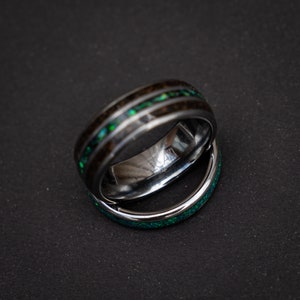 May include: Two rings, one with a black and green wood inlay and the other with a green opal inlay. The rings are stacked on top of each other.