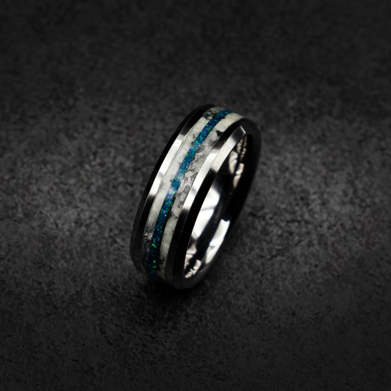 May include: A black tungsten carbide ring with a white and blue inlay. The inlay is made of opal and has a speckled pattern.