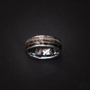 Op de afbeelding: Een zilveren ring met een zwarte en gouden inlay. De inlay is gemaakt van een materiaal dat eruitziet als gebroken steen.