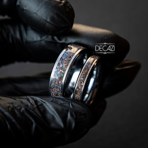 May include: Two silver rings with a brown and blue speckled inlay. The rings are shown together in a black gloved hand. The text "DECAZI" is visible in the background.