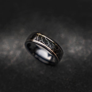 May include: A black tungsten carbide ring with a gold inlay featuring a swirling pattern.