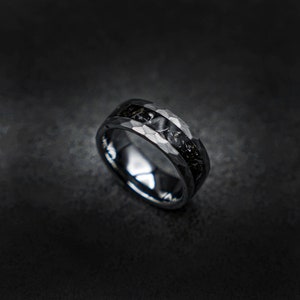 Op de afbeelding: Een donkere, gehamerde metalen ring met een zwarte inleg. De ring heeft een getextureerd, geometrisch patroon op de buitenste band en een donker, mogelijk steenachtig, midden. De ring is geplaatst tegen een donkere, gevlekte achtergrond.