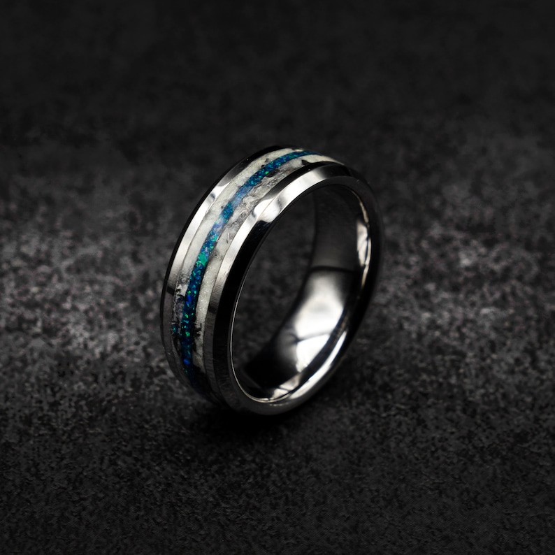 May include: A silver tungsten carbide ring with a blue and white inlay. The ring has a bevelled edge and a polished finish.
