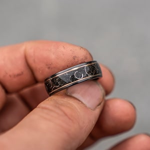 May include: A black ring with a gold inlay design. The ring has a textured surface and is worn on a finger.