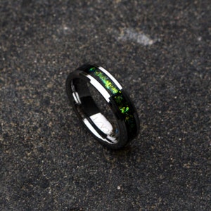 Op de afbeelding: Een zwarte ring van wolfraamcarbide met een groene en zilveren inlay. De ring heeft een afgeschuinde rand en een gepolijste afwerking.