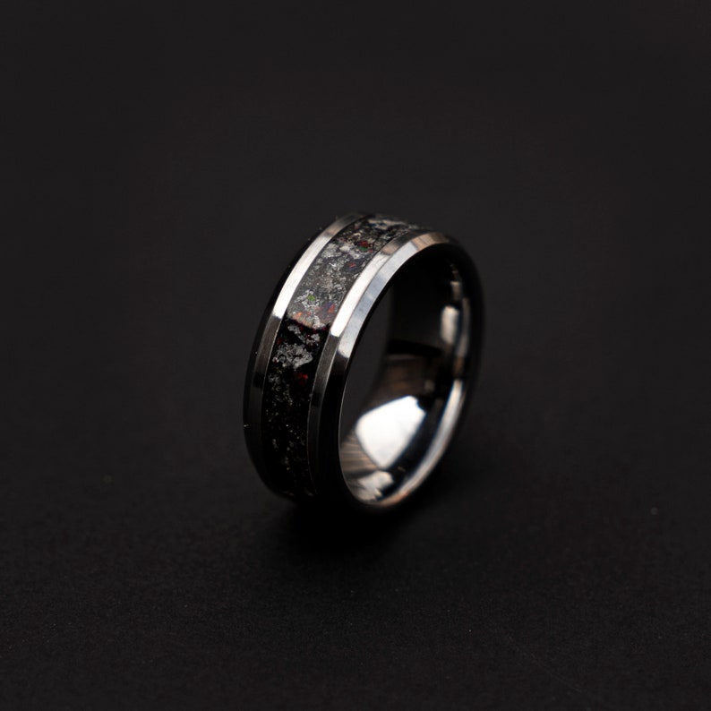 May include: A black and silver tungsten carbide ring with a black inlay. The inlay has a speckled pattern of silver and red.