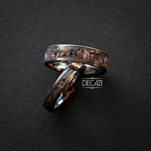 May include: Two silver rings with a black inlay and blue and brown flecks. The rings are stacked on top of each other.