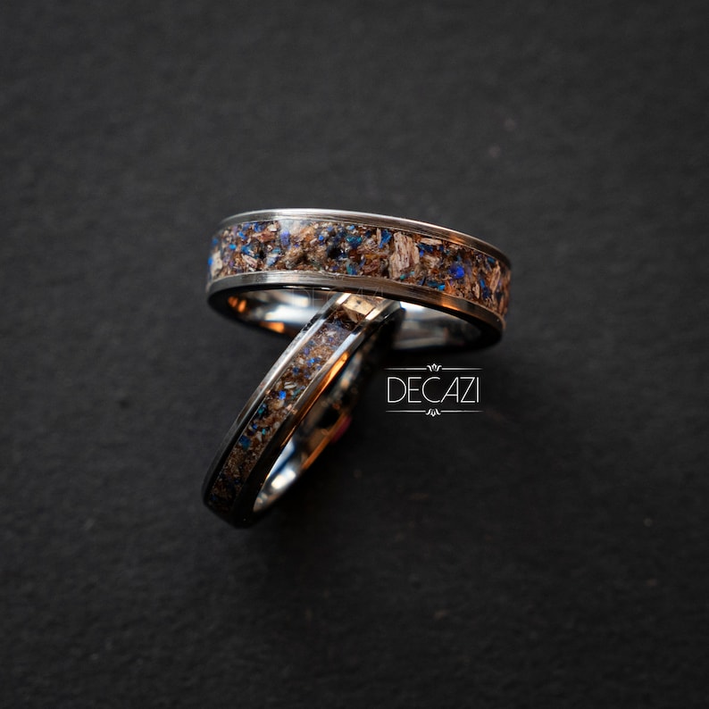 May include: Two silver rings with a black inlay and blue and brown flecks. The rings are stacked on top of each other.