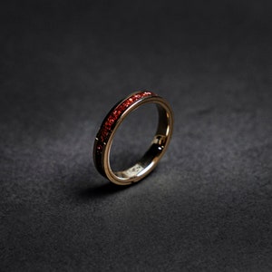 Op de afbeelding: Een goudkleurige ring met een band van kleine, rode edelstenen. De ring staat tegen een donkere achtergrond, wat het contrast tussen het goud en de rode stenen benadrukt. Het ontwerp van de ring is eenvoudig en elegant.