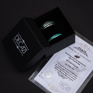 May include: Two rings in a black box. The rings are made of metal and have a green and black inlay. The box has a white label with the text "DECAZI" and a certificate of authenticity for a Megalodon fossil. The certificate states that the fossil was found in South Carolina, USA and is 15 million years old.