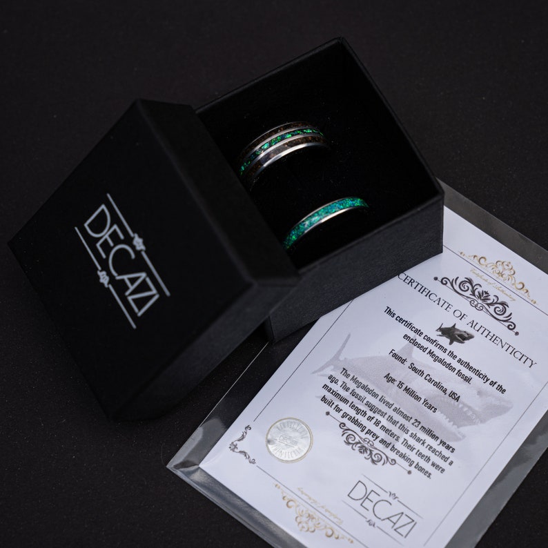 May include: Two rings in a black box. The rings are made of metal and have a green and black inlay. The box has a white label with the text "DECAZI" and a certificate of authenticity for a Megalodon fossil. The certificate states that the fossil was found in South Carolina, USA and is 15 million years old.