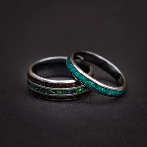 May include: Two silver rings with green and black inlay. The ring on the left has two thin green lines and a black inlay. The ring on the right has a single green line.