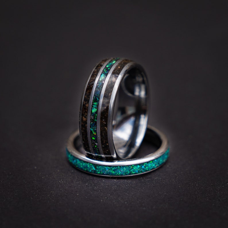 May include: Two silver rings with a black and green inlay. The top ring has a wider band and a green opal inlay. The bottom ring has a thinner band and a green opal inlay.