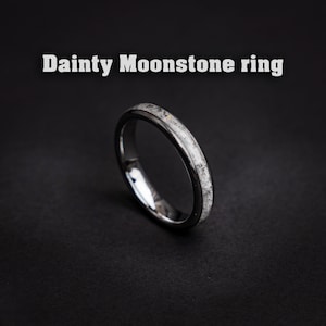 Può includere: Un anello d'argento delicato con una fascia sottile e un'incisione di pietra lunare. L'anello è su uno sfondo nero.