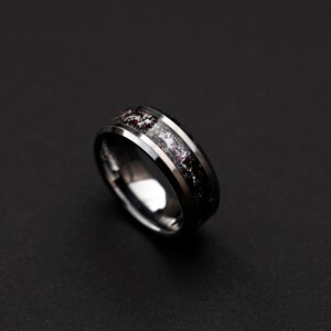 May include: A black tungsten carbide ring with a beveled edge and a black and white inlay. The inlay has a speckled pattern of white and red.