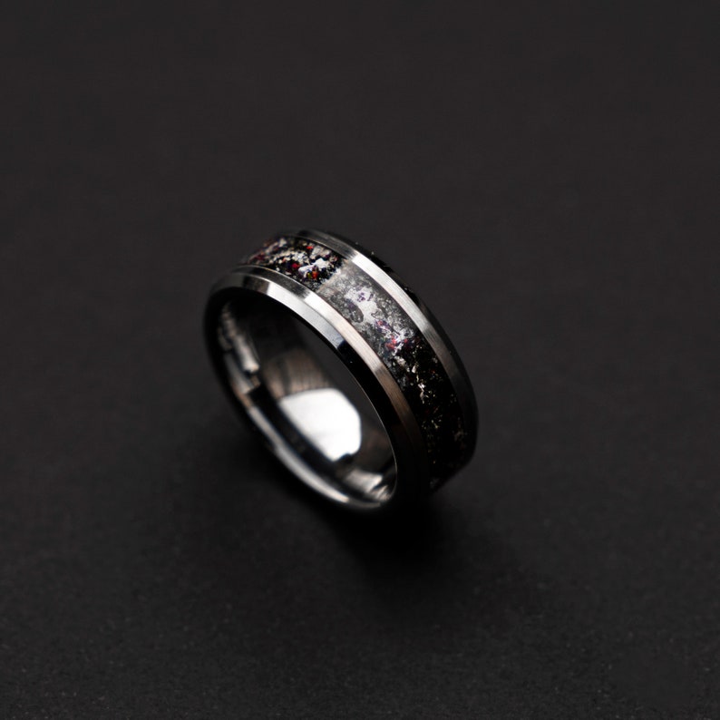 May include: A black tungsten carbide ring with a beveled edge and a black and white inlay. The inlay has a speckled pattern of white and red.