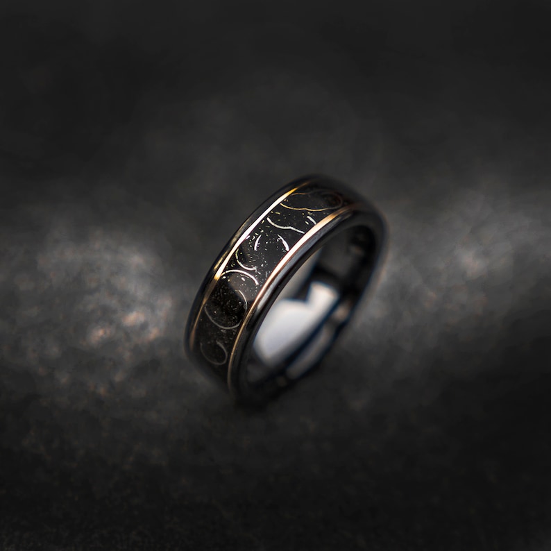 May include: A black tungsten carbide ring with a gold-colored band and a swirling design. The ring is on a dark gray surface.