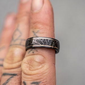 May include: A black and gold ring with a textured design. The ring is worn on a finger.